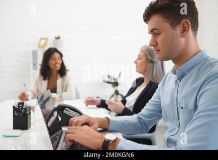 Young focused businessman typing on his laptop in an office at work. Mature caucasian businesswoman talking to an african american colleague while a Stock Photo