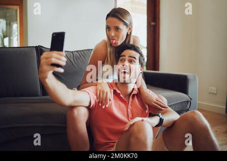 Funny romantic couple pulling faces and fooling around while holding a mobile phone and taking a selfie together. Playful man and woman sitting Stock Photo