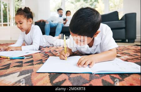 Little boy and girl drawing with colouring pencils lying on living room floor with their parents relaxing on couch. Little children sister and brother Stock Photo