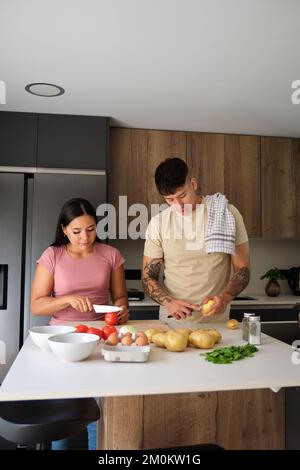 Two young latin people peeling potatoes and cutting tomatoes at kitchen. Stock Photo