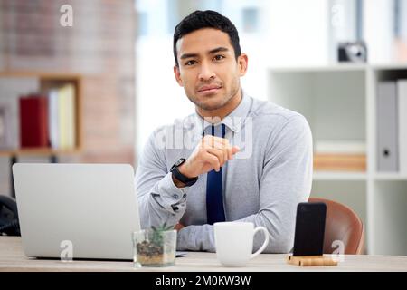 Young serious mixed race businessman working on a laptop alone in an office at work. One focused confident hispanic businessperson sitting at a desk Stock Photo
