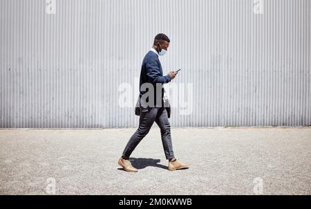 Businessman wearing face mask while commuting during the covid pandemic. Man using cellphone while walking to work. Using internet while walking in Stock Photo