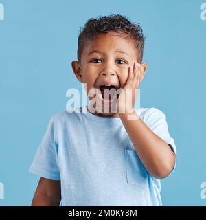 Cute hispanic little boy with hand on face and mouth open being surprised and shocked showing true astonished reaction against a blue studio Stock Photo