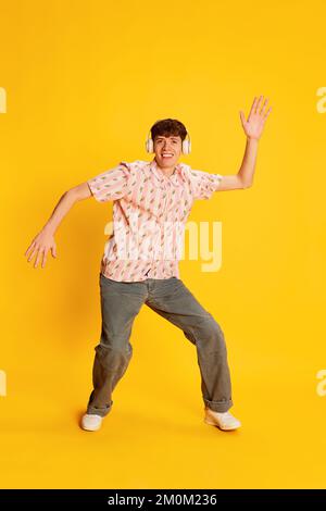 Excited young man, happy dude in vintage style clothes listening to music and dancing in headphones isolated over bright yellow background. Stock Photo