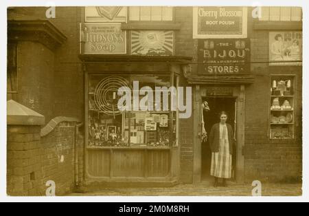 Original early1920's postcard of proprietor of M. Humphries, toys and glassware shop, also sign advertises 'the Bijou hardware  & china stores'.  Perhaps this is a franchise operation. The woman in the doorway, who is probably the owner, looks drab, her dress hangs loosely, ill-fitting and shapeless, a housedress type - it has checks fashionable at this time. Her hair is tied in a bun at the back. She looks poor. No sign of the flapper decadence of the era here! Circa 1923, U.K. Stock Photo