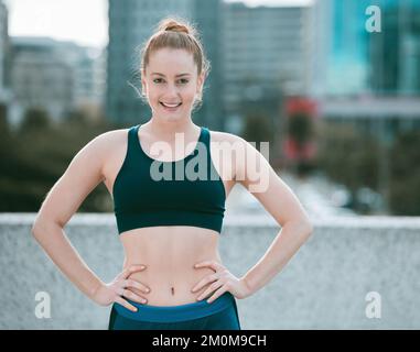 Portrait of one confident young caucasian woman standing with hands on hips ready for exercise outdoors. Determined female athlete looking happy and Stock Photo