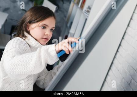 Close-up view of girl, child is wiping off the white board in class with sponge. Kid erases something written on chalkboard. Childhood concept. Educat Stock Photo