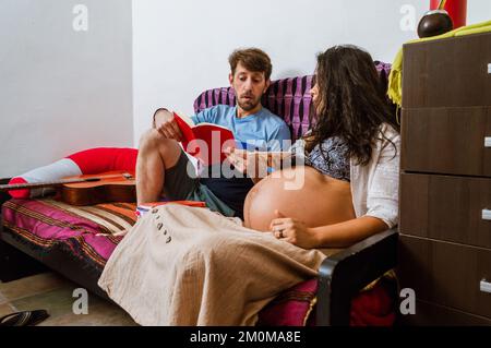 caucasian adult man with beard, he is concentrated sitting reading a story in a book to his pregnant wife indoors, sitting in the living room, lifesty Stock Photo