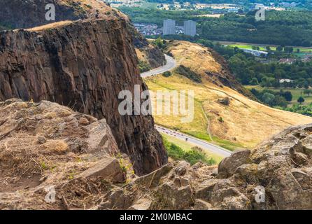 Visitors climbing to Arthur's seat,stand at the precipice of a high vertical cliff in awe of views across Edinburgh City,on a summer day in Scotland's Stock Photo