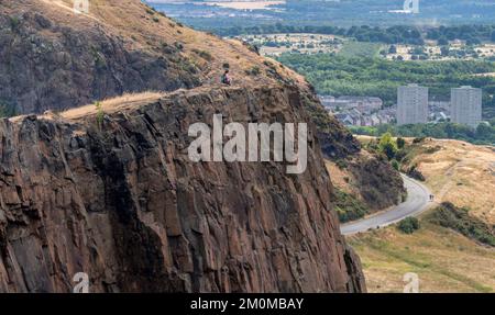 Visitors climbing to Arthur's seat,stand at the precipice of a high vertical cliff in awe of views across Edinburgh City,on a summer day in Scotland's Stock Photo