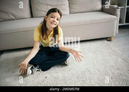 high angle view of cheerful girl sitting on floor with crossed legs and open palms near couch at home Stock Photo