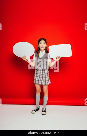 full length of girl in checkered skirt and gumshoes holding blank speech bubbles on red background Stock Photo