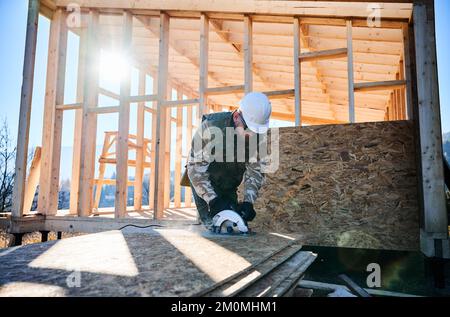 Carpenter using circular saw for cutting wooden OSB board. Man worker building wooden frame house on pile foundation. Carpentry concept. Stock Photo