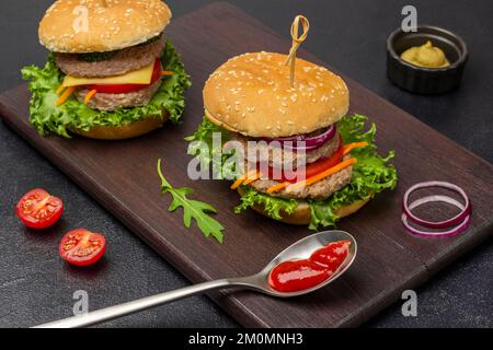 Two beef burgers with greens, chopped onions and carrots on board. Tomato sauce in spoon. Black background. Stock Photo