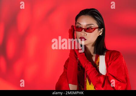 trendy asian woman in red gloves and trendy sunglasses holding hands near face on abstract red background,stock image Stock Photo