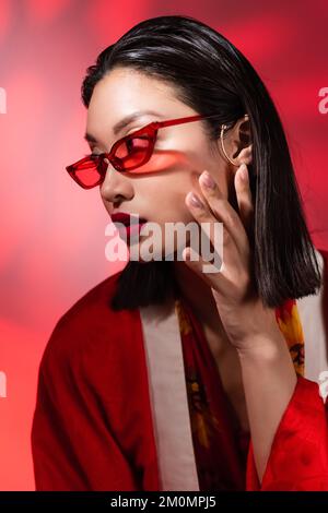 asian woman in trendy sunglasses and ear cuff posing with hand near face on abstract background with red gradient,stock image Stock Photo