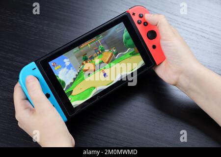 Defocused view to girl playing Super Mario Odyssey game on Nintendo Switch console in handheld mode Stock Photo