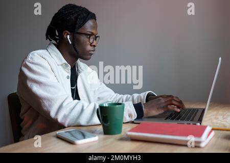 Young focused African American man sits at wooden table with laptop getting internet education Stock Photo