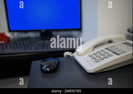 Desk with a black mouse and white telephone with a monitor screen on blue on the background and a keyboard also. Shallow depth of field Stock Photo