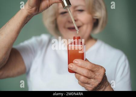 Close-up portrait older female's hands holding red bottle with facial essence dropping collagen moisturizer face serum. Rejuvenation and skincare Stock Photo