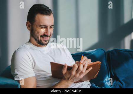 Smiling young student man wearing white t-shirt sitting on blue sofa indoors writing down memories in notebook diary, resting on weekends at home Stock Photo