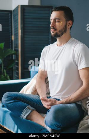 Handsome man practicing meditation in the cross-legged posture, on the sofa in his living room. Breathing exercise. Stock Photo