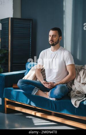 Man sitting with his legs crossed on a sofa and meditating in living room. Concept of supporting developmental health and brain development. Stock Photo