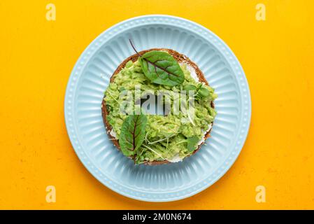 Bagel with cream cheese, avocado and micro greens on a blue plate, yellow background. Top view. Healthy vegetarian snack food Stock Photo
