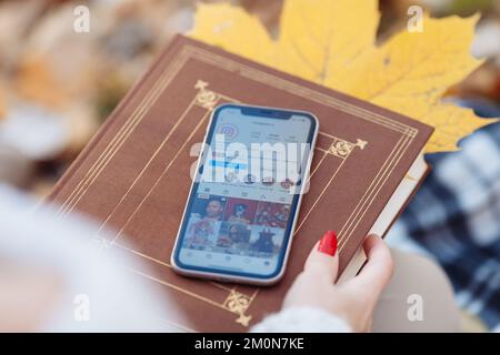 the Instagram page is open on the iPhone 11, which the girl is holding on the street with a book in autumn Stock Photo