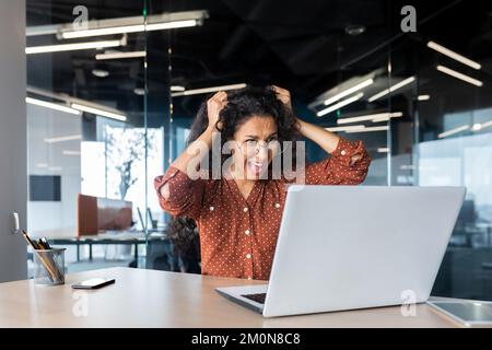 Upset woman at work yelling at laptop monitor, angry businesswoman holding hair on head inside office. Stock Photo