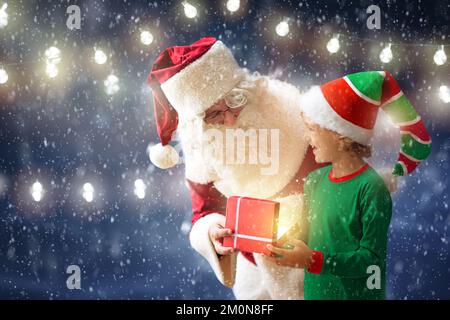 Santa and elf deliver Christmas presents. Santa Claus and little child open a gift box with magical glow. Fairy lights in in snowy forest. Stock Photo