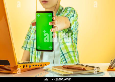 Homeschooling concept and using laptop study online. School boy in headphones holding phone with green screen have video call distant at class distanc Stock Photo