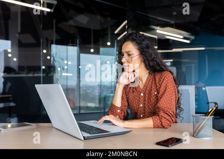 Serious thinking Latin American woman working inside office, businesswoman pondering complex decisions using laptop at work. Stock Photo