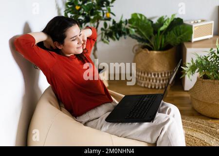 Relaxed young woman sitting in a bag chair with stretched arms using a laptop surrounded by artificial plants Stock Photo