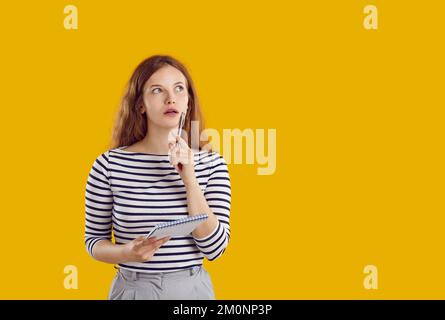 Woman in striped shirt is thinking holding pen and notebook looking up on yellow background. Stock Photo