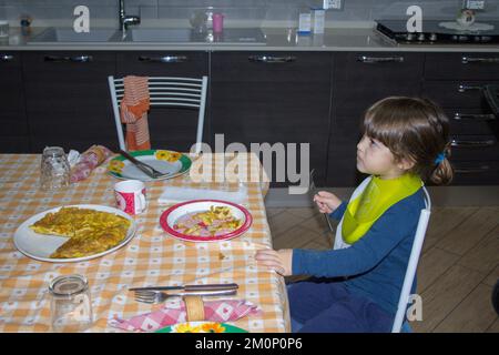 Image of an adorable little brunette girl wearing bibs, sitting at the table eating an omelette and enthralled watching tv. Distraction of children Stock Photo