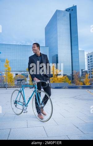 Young smiling stylish businessman going to the office by bicycle in front of financial district. Stock Photo