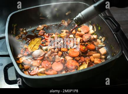 Red wine is poured into a cooking pan of steaming roasted vegetables to deglaze for a sauce base from carrots, onions, leeks, celery and bay leaves, c Stock Photo
