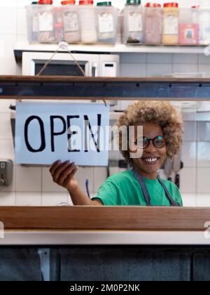 Happy African American woman looking at camera and Hanging Open Sign by the Glass Window Stock Photo