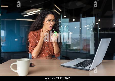 Sick woman in the office, Latin American business woman has a severe sore throat, uses medicine to relieve the pain. Stock Photo