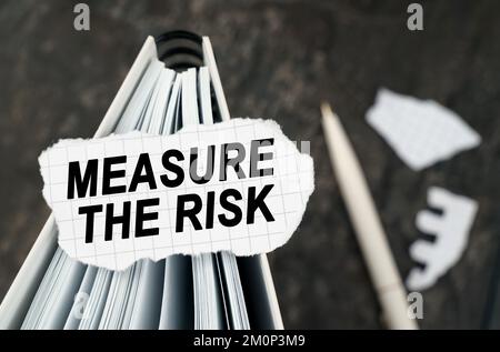 On the table is a notebook on which lies a piece of torn paper with the inscription - Measure The Risk. The pen lies outside the sharpness zone. Stock Photo