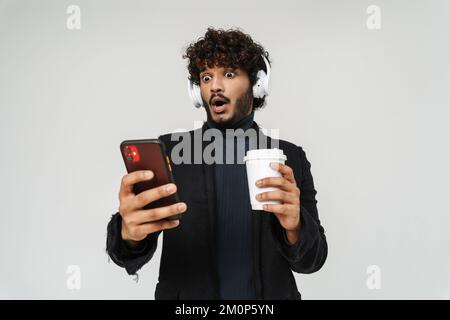 Young handsome curly surprised indian man in headphones with coffee and phone over isolated grey background Stock Photo