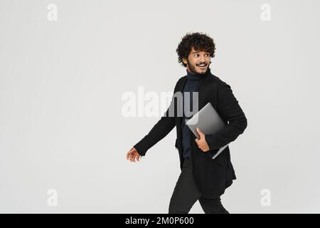 Young handsome curly smiling indian man walking with laptop over isolated grey background Stock Photo