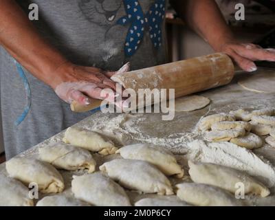 hands of a mature woman roll a wooden rolling pin over a piece of dough on the kitchen table and shaped burgers in the foreground in defocus Stock Photo