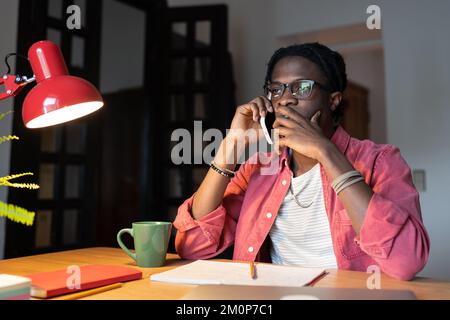 Worried frustrated African student guy making call, talking on mobile phone while studying at home Stock Photo