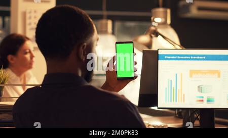 Startup employee holding smartphone with greenscreen and working on company data, looking at isolated mockup display. Using blank chroma key copyspace on mobile phone template. Stock Photo