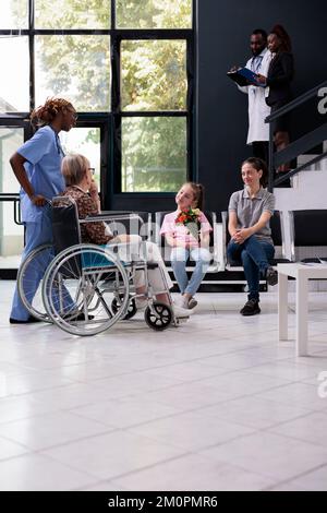 Nurse bringing elderly patient to family after finishing medical appointment while explaining health care treatment in hospital waiting area. Granddaughter holding bouquet of flowers for grandmother Stock Photo