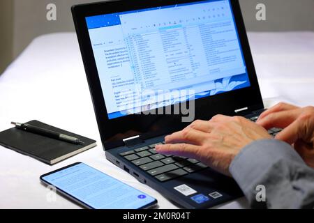 A closeup of a person's hands typing into a laptop computer with a notebook and smartphone nearby. Stock Photo