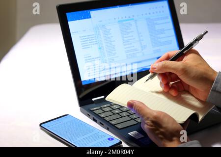 A closeup of a person's hands holding a pencil and notebook writing down information from documents displayed on a laptop computer. Stock Photo