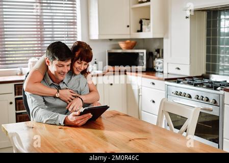 This is the app I told you about last night. a man using a digital tablet while being embraced by his wife at home. Stock Photo